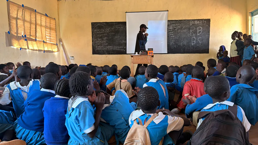 Pupils in blue uniforms seated on a classroom floor, facing a projector screen and blackboard during a demonstration.