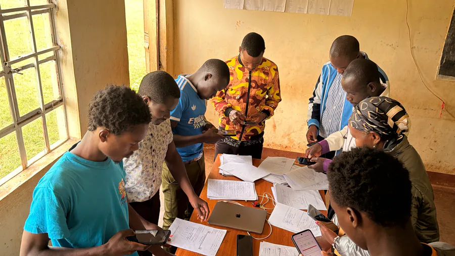 Teachers standing around a classroom table covered in examination scripts, each working through papers on a phone.